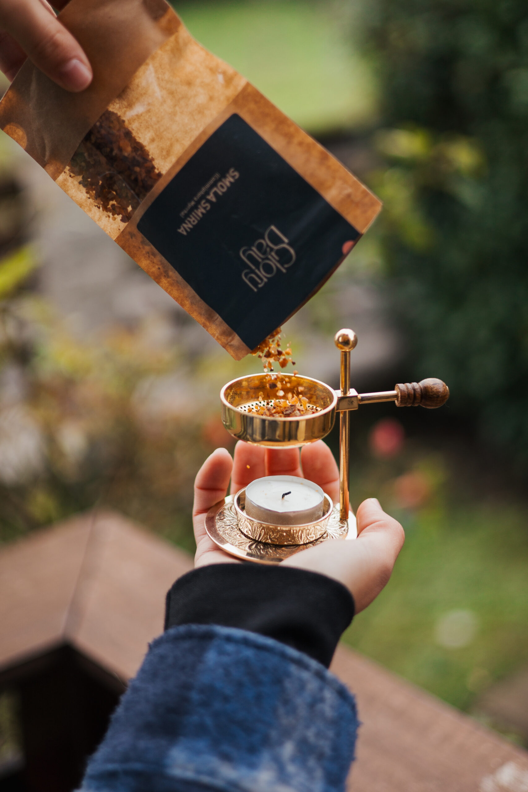 Osoba sipa aromatičnu smjesu iz smeđe vrećice brenda Glory day na metalnu aroma lampu s upaljenom svijećom, na otvorenom prostoru s nejasnom pozadinom prirode.