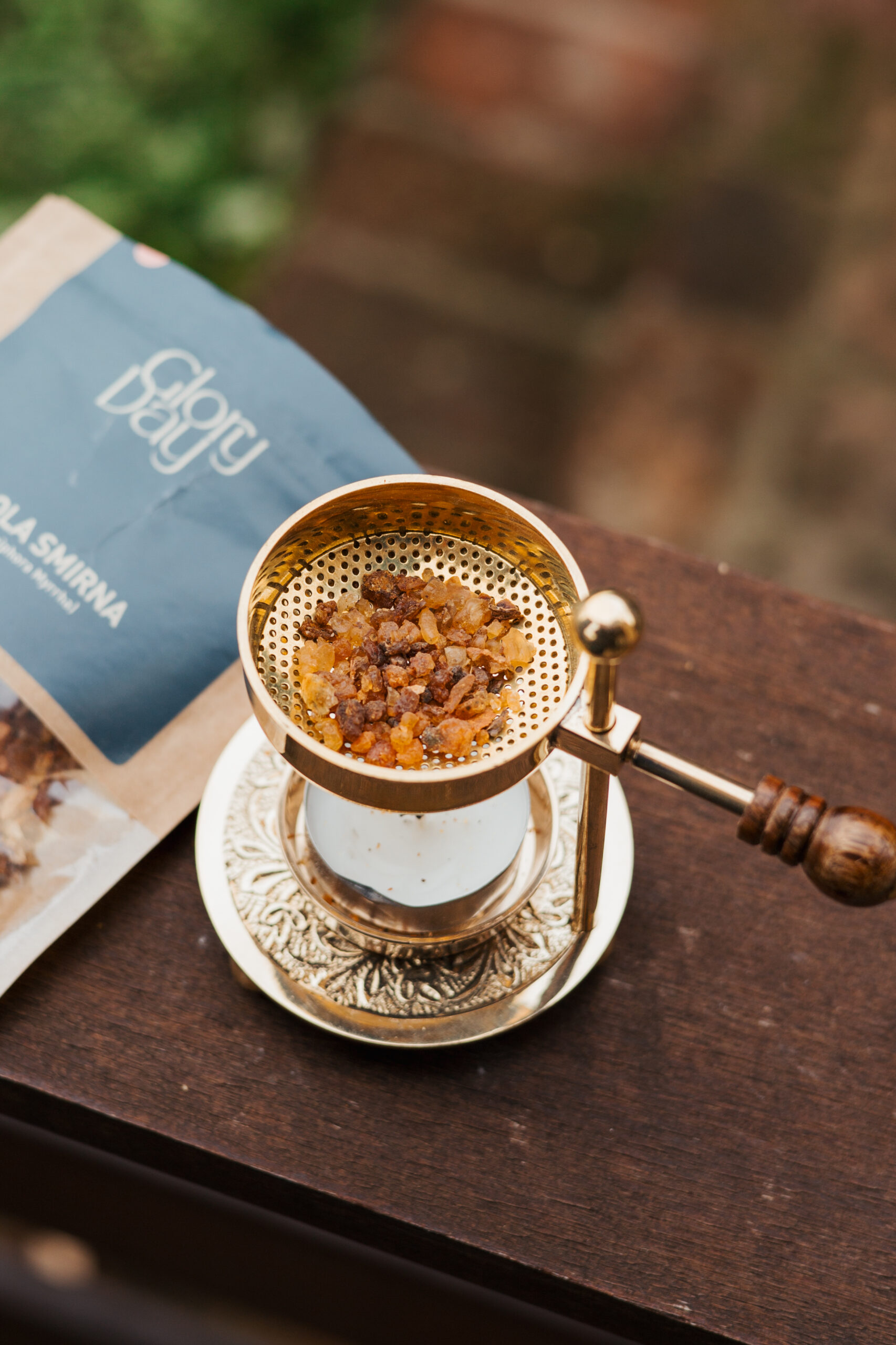 Zlatna metalna kadilica s mirisnim smolama na drvenoj površini pored paketa tamjana s natpisom 'Glory Day'.