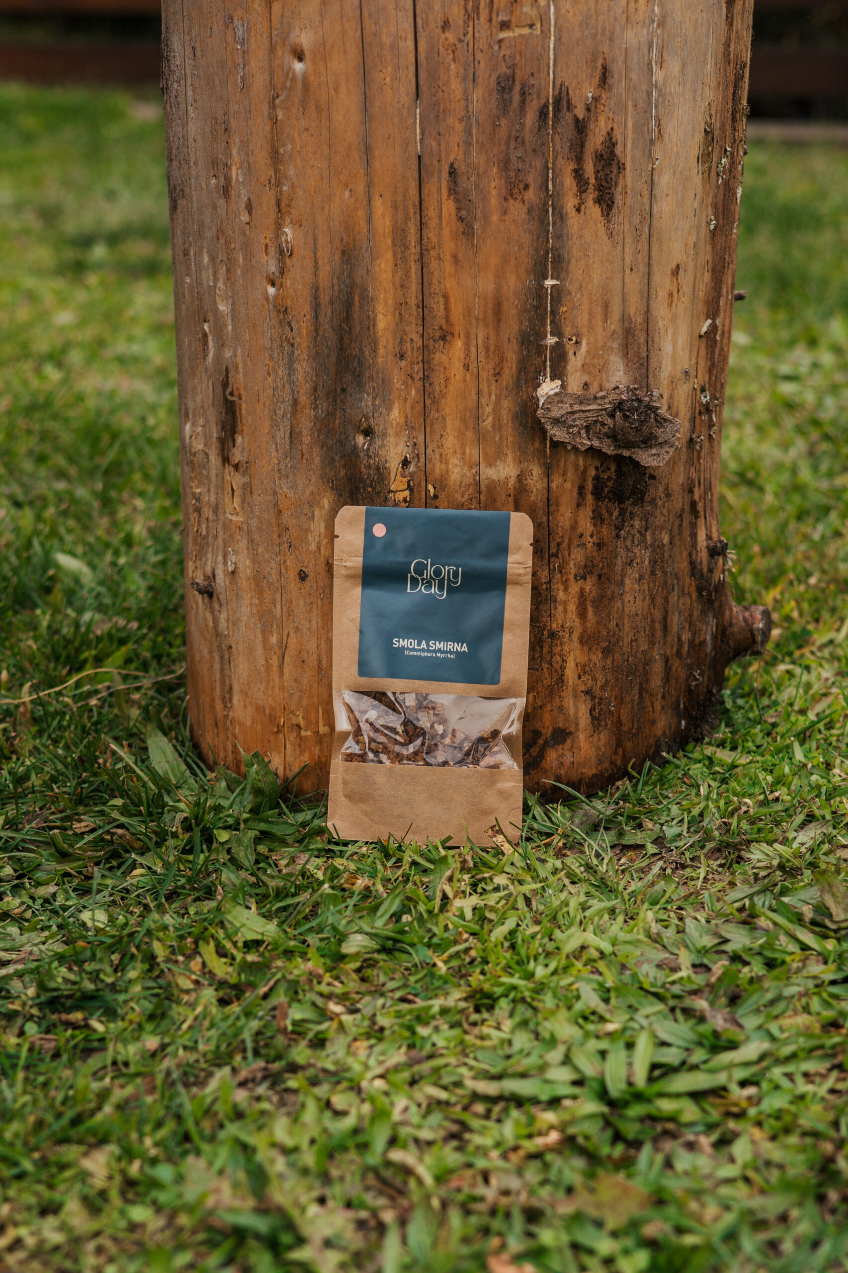 Smeđi paketić smole smirne s etiketom "Glory Day" naslonjen na deblo drveta na travnatoj površini.