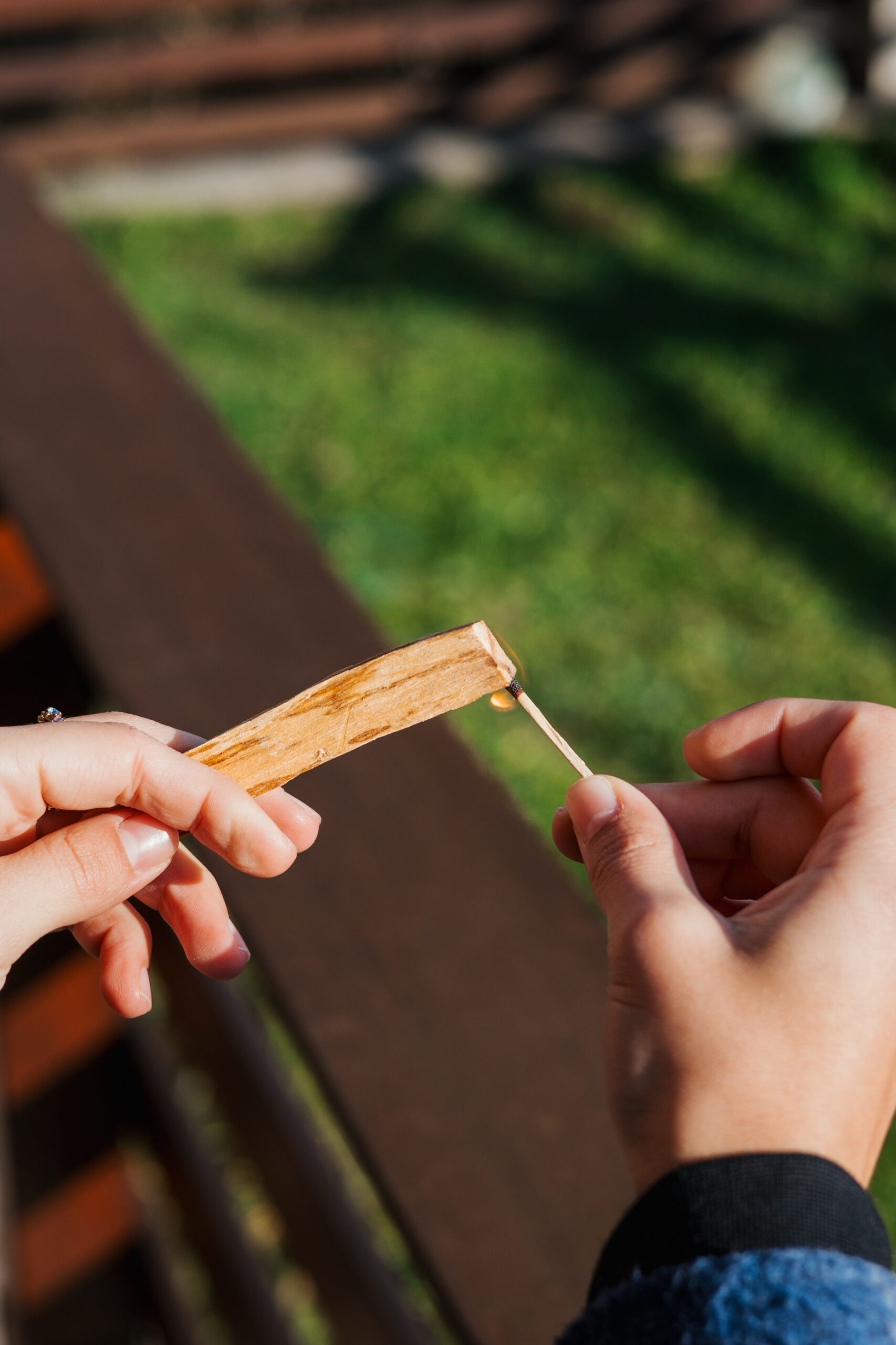 Osoba drži štapić palosanto drva u jednoj ruci, dok drugom rukom pali drvo šibicom, na otvorenom prostoru s travom u pozadini.