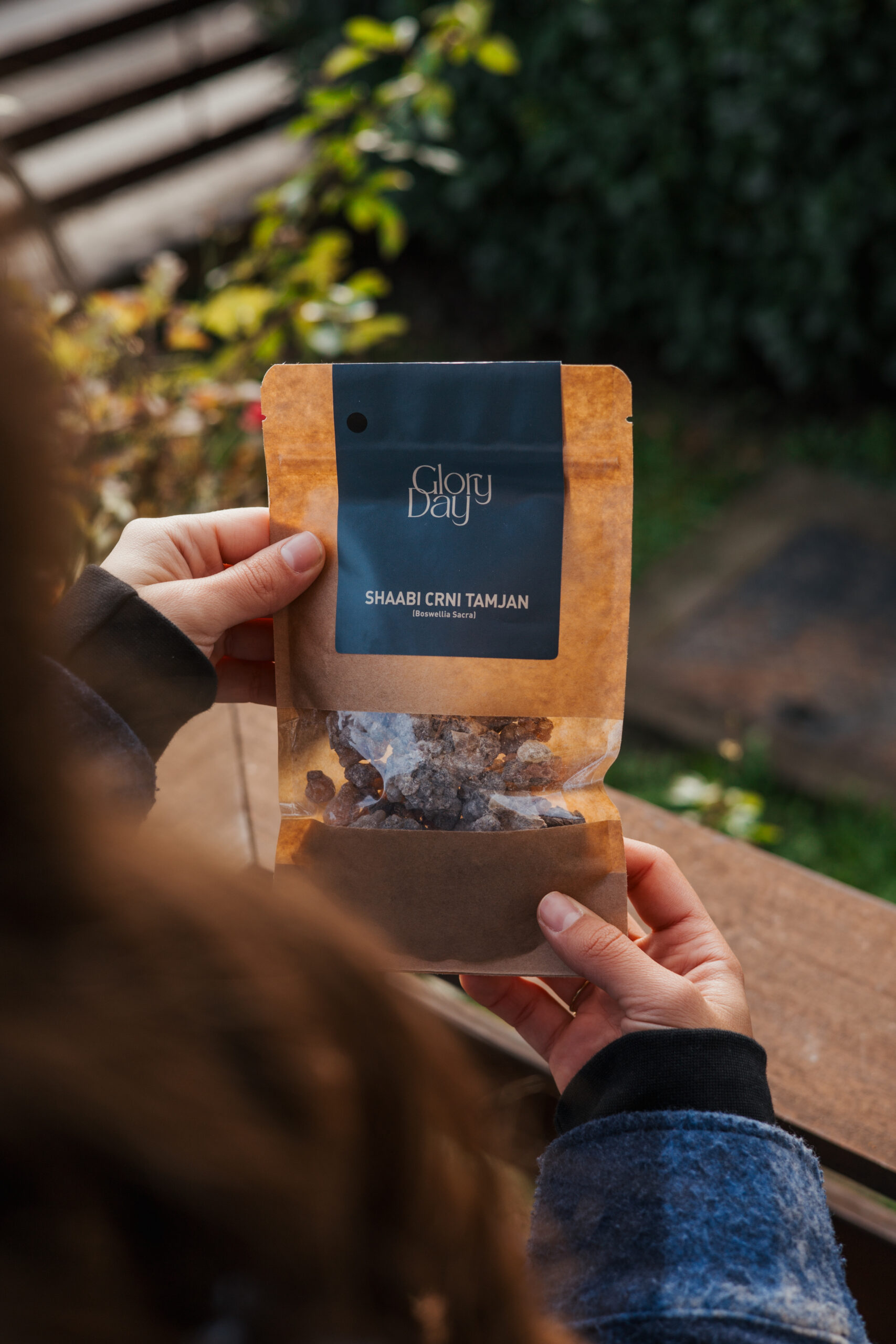 Osoba drži pakiranje tamjana s natpisom 'Glory Day: Shaabi Crni Tamjan (Boswellia Sacra)', snimljeno na otvorenom s biljkama u pozadini.