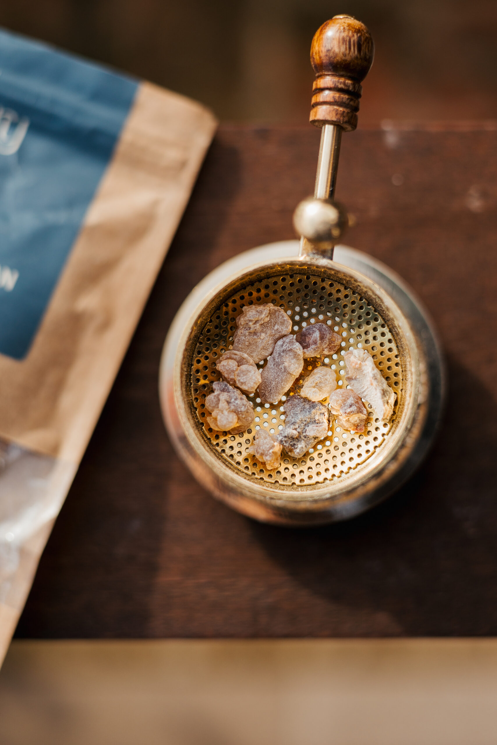 Metalna kadilica s komadićima smole, postavljena na drvenu površinu uz pakiranje sa strane.