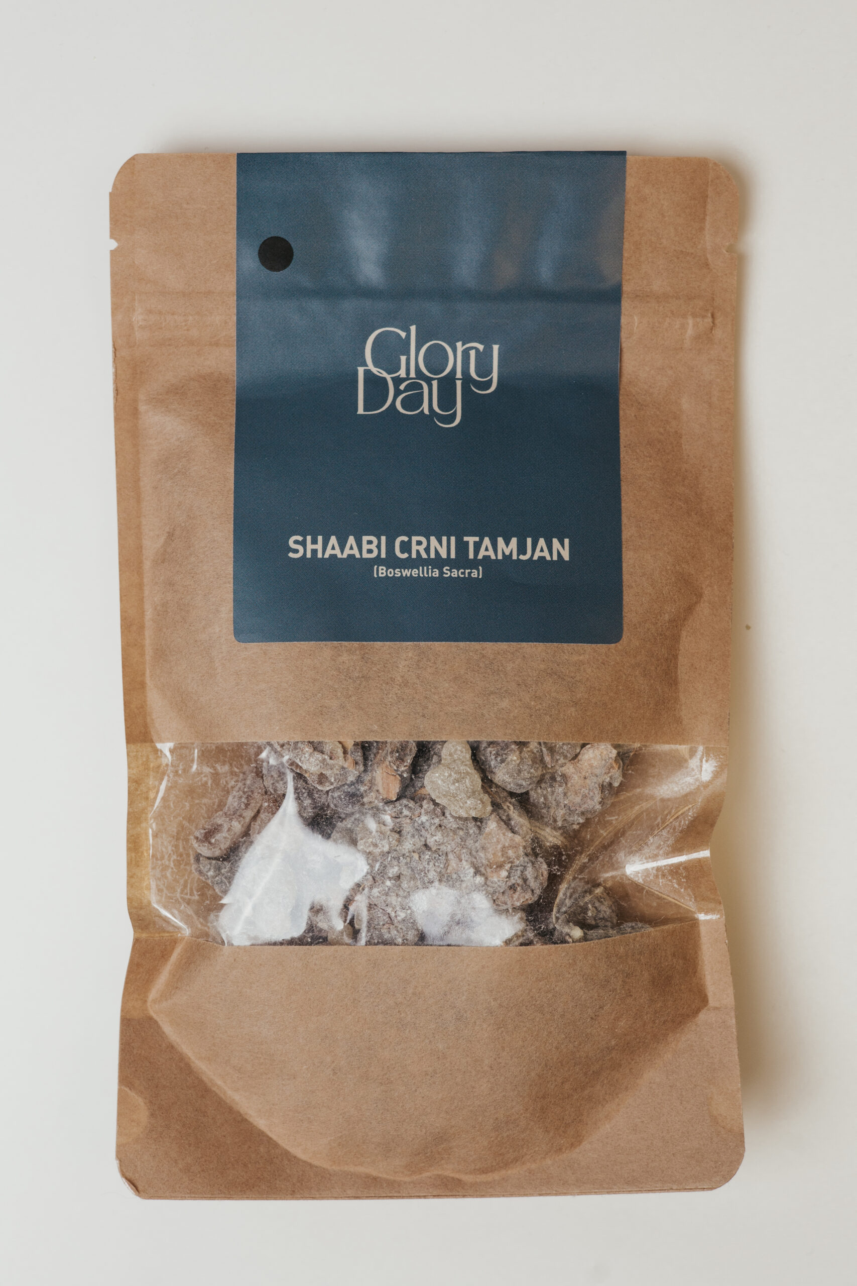 Pakiranje smeđeg papira s plavom etiketom na kojoj piše 'Glory Day' i 'Shaabi Crni Tamjan (Boswellia Sacra)' s prozirnim dijelom kroz koji se vidi unutrašnji sadržaj tamjana.