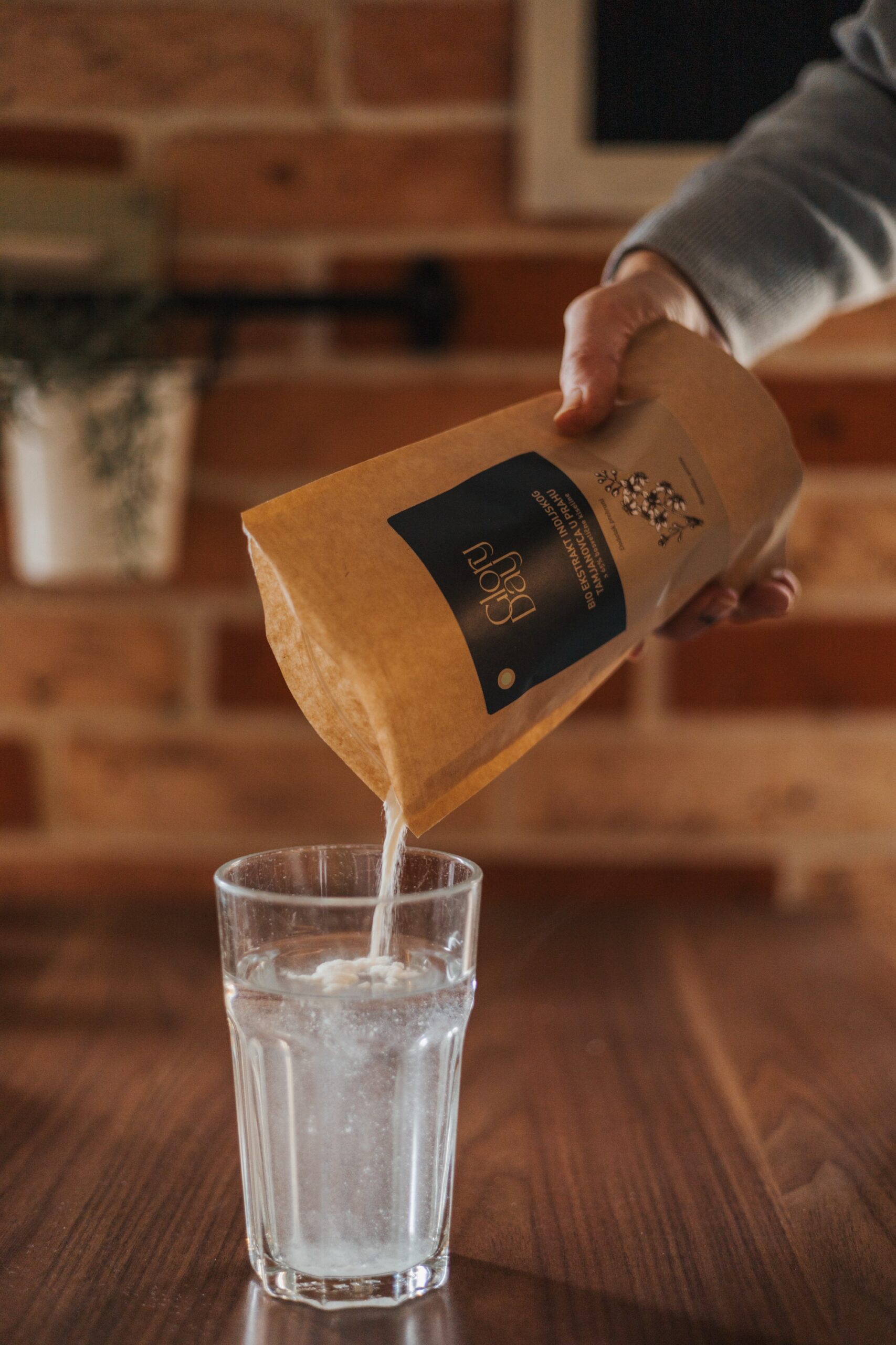 Čaša s vodom na drvenom stolu dok osoba sipa prašak iz smeđe papirnate vrećice brenda Glory day, s etiketom na kojoj je crni logotip i ilustracija biljke, u čašu.